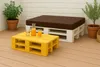 An outdoor seating area with a yellow pallet coffee table and a white pallet bench with a brown cushion. There are two glasses of orange juice and a bottle of orange juice on the coffee table. Two potted plants are visible in the background. Produktbild-Vorschau