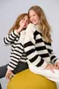 Two young girls wearing matching striped sweaters, one whispering to the other, sitting on a yellow pouf. Produktbild-Vorschau