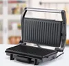 An open electric grill on a white surface with a blurred kitchen background. Produktbild-Vorschau