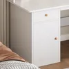 A close-up view of a white desk with a drawer and a cabinet door, featuring wooden knobs. The desk is positioned next to a bed with a brown pillow and a patterned blanket. Podgląd zdjęcia produktu