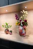 Two small potted plants and a vase of flowers are displayed on a wooden shelf. Produktbild-Vorschau