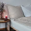 A bed with pillows and a duvet, a bedside table with a candle and flowers. Produktbild-Vorschau