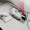 A digital alarm clock with a red projector beam is placed on a desk next to an open book. Náhled obrázku produktu