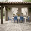 An outdoor dining set with blue chairs and a white table on a stone patio, under a tiled roof. Produktbild-Vorschau
