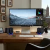 A modern desk setup with a computer monitor, keyboard, speakers, and framed pictures on the wall, with a city view in the background. Náhľad obrázku produktu