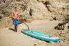 A man is pumping up a paddleboard on a sandy beach with rocky cliffs in the background. Produktbild-Vorschau