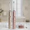Two AILORIA electric toothbrushes, one gold and one rose gold, are displayed on a marble surface in front of a blurred bathroom background. A small charging base is also visible. Náhled obrázku produktu