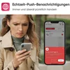 A woman is looking at her phone, which displays a smoke alarm notification. The background is blurred. Produktbild-Vorschau