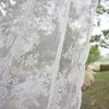A close-up view of a white lace curtain with floral embroidery, with a blurred outdoor scene in the background. Produktbild-Vorschau