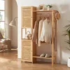 A wooden wardrobe stand with clothes hanging on it, a hat and scarf on the side, and a handbag on a shelf. The background shows a room with a window and a lamp. Produktbild-Vorschau