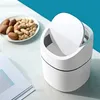 A small white trash can with its lid open, placed on a white surface next to a plate of peanuts and a book. Produktbild-Vorschau