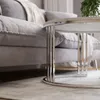 A close-up view of a round coffee table with a silver chrome base and a white top, placed in front of a gray sofa with a beige throw blanket. Náhľad obrázku produktu