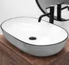 A white oval basin with a black rim sits on a wooden surface, with a black faucet and a round mirror in the background. Anteprima dell’immagine prodotto