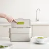 A hand is shown pressing a green button on the lid of a compost bin, which is placed on a kitchen counter next to a sink and a bowl of edamame. Náhled obrázku produktu