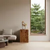 A wooden decorative column with a sculpture on top, placed in a living room next to a sofa, with a large window showing a garden view. Voir les images du produit
