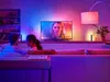 A couple sits on a couch in a living room, watching TV. The room is illuminated by colorful ambient lighting from LED light strips behind the TV and a floor lamp. Anteprima dell’immagine prodotto