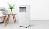 A white portable air conditioner is shown in a living room setting next to a small wooden table with a plant and books. Produktbild-Vorschau