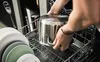A person is loading a pot into a dishwasher rack, with plates stacked to the left. Anteprima dell’immagine prodotto