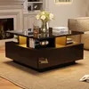 A modern living room scene featuring a black high-gloss coffee table with gold accents and integrated lighting. The table is set with books, a glass of liquid, and a vase of flowers. A fireplace and a sofa are visible in the background. Náhľad obrázku produktu