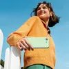 A young woman is holding a green smartphone case while leaning on a white fence with a blue sky in the background. Produktbild-Vorschau