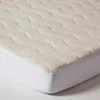 A close-up, angled view of a cream-colored fleece mattress topper with a textured, wavy pattern, placed over a white fitted sheet. Anteprima dell’immagine prodotto