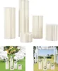 A collection of white, fluted cylindrical pedestals of varying heights, displayed against a white background. Below this, two lifestyle shots show the pedestals used as decorative elements in event settings, one with balloons and signage, the other with fl Podgląd zdjęcia produktu