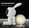 A white astronaut rabbit figurine sits on a moon-like surface next to a glowing moon sphere, with a starry night sky in the background. Náhled obrázku produktu
