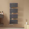 A dark gray towel radiator mounted on a light brown wall, with bathroom accessories on a shelf to the left and a small vase with dried grass on a wooden block to the right. Náhľad obrázku produktu