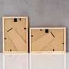 The backs of two wooden picture frames are shown against a gray concrete wall and a white reflective surface. Produktbild-Vorschau