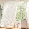 A bright room with a large window covered by sheer curtains with a cloud pattern. A small cabinet and framed pictures are visible. Produktbild-Vorschau