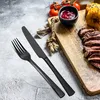 A black cutlery set is laid out on a textured gray surface next to a wooden cutting board with sliced steak, corn on the cob, and a small bowl of sauce. Produktbild-Vorschau