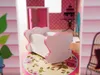 A close-up view of a dollhouse crib on a floral patterned surface, with other dollhouse furniture and decor visible in the background. Voir les images du produit