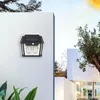 A solar-powered outdoor light mounted on a white wall, with a modern building and trees in the background under a sunny sky. Produktbild-Vorschau