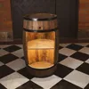 A wooden barrel-shaped cabinet with shelves, standing on a black and white checkered floor in front of a brick wall and a dark door. Náhled obrázku produktu