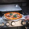 A pizza is being placed on a grill using a pizza peel, with a pizza oven attachment visible. Podgląd zdjęcia produktu