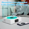 A person is sitting on a couch wrapped in a blanket, reading a book, while a Bluetooth vinyl record player with a water wave light is in the foreground. Produktbild-Vorschau