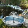 A solar-powered fountain kit is operating in a galvanized metal basin placed next to a pond. Produktbild-Vorschau