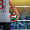 A person holding a volleyball in front of a blurred volleyball net and stadium seating. Voir les images du produit