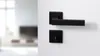 A black door handle and lock set mounted on a white wall, with a blurred interior scene in the background. Produktbild-Vorschau