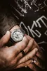 A close-up shot of a person's hands wearing a silver watch and rings, with a dark, graffiti-covered background. Náhled obrázku produktu