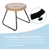 A small round table with a glass top and woven rattan edge, supported by black metal legs. A close-up inset shows an adjustable foot on one of the legs. Text describes the adjustable feet. Produktbild-Vorschau