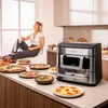 A woman is relaxing by a window in a kitchen, with a counter full of food and an air fryer. Produktbild-Vorschau