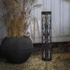 A black solar-powered pathway light with a decorative, branch-like pattern stands on a gravel surface next to a large, dark, textured planter filled with dried grass. Produktbild-Vorschau