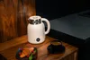 A white electric kettle is steaming on a wooden surface next to a cup of coffee and a croissant. Podgląd zdjęcia produktu