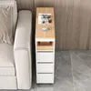A narrow cabinet with drawers and a shelf sits next to a sofa, with a magazine and coffee cup on top. Produktbild-Vorschau