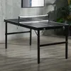 A black foldable mini ping pong table with a net is set up on a gray floor in front of a concrete wall. Podgląd zdjęcia produktu