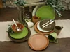 A collection of stoneware dishes and mugs arranged on a table with a rustic wooden crate and olive branches in the background. Náhľad obrázku produktu