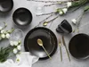 A table setting with black plates, gold cutlery, a black mug, and white flowers. Produktbild-Vorschau