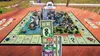 A 3D Monopoly board game set up on a wooden table, with miniature city buildings and water features. Cards and money are scattered around the board. Produktbild-Vorschau