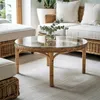 A round glass-top coffee table with a rattan base is placed on a rug in a living room setting, with a sofa and cushions in the background. Produktbild-Vorschau